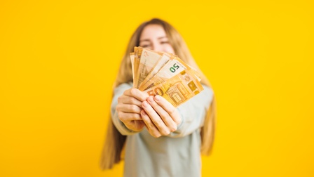 Eine Person steht vor einem gelben Hintergrund und hält einige 50-Euro-Geldscheine in die Kamera.