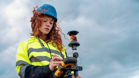 Person mit Helm in Schutzbekleidung steht unter blauem Himmel, Wind weht durch Haar und blickt auf Messgerät, das sie in Händen hält.