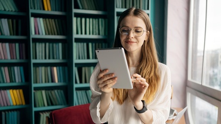 Jugendliche Person mit langen Haaren, Brille und weißer Bluse hält ein Tablet in Händen, im Hintergrund steht eine Couch sowie Regale mit Büchern vor einer offenen hellen Fensterfront