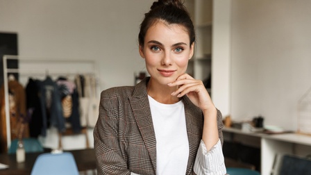 Porträt einer Person mit dunklen geschlossenen Haaren, weißem Shirt und kariertem Blazer in einem Büro