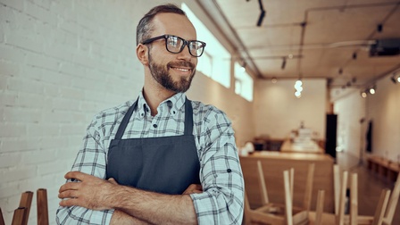 Lächelnde bärtige Person mit Brillen in dunkelblauer Schürze hält Arme verschränkt vor Körper und blickt zur Seite im Hintergrund verschwommen Raum mit weißen Ziegelwänden mit Holztischen auf denen umgedrehte Stühle gestellt sind und Holztheke 
