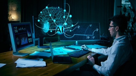 Eine Person sitzt an einem Schreibtisch und arbeitet an mehreren Bildschirmen. Auf den Monitoren sind grafische Darstellungen und Benutzeroberflächen zu sehen. Über den Bildschirmen ist eine leuchtende Netzwerkgrafik mit dem Schriftzug „AI“ eingeblendet