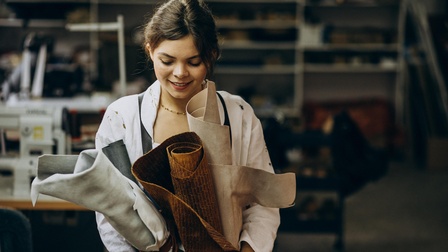 Eine schräg nach unten blickende Person hält mit beiden Händen vier große, unterschiedliche Stoffrollen. Hinter ihr steht eine Nähmaschine