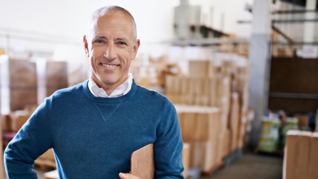 Person mit Pullover steht mit einem Klemmbrett und Kugelschreiber freudig in einer Lagerhalle