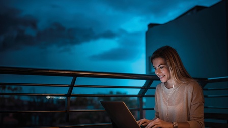 Eine junge Frau am Laptop auf einer Dachterrasse. Blaue Dämmerung im Hintergrund.