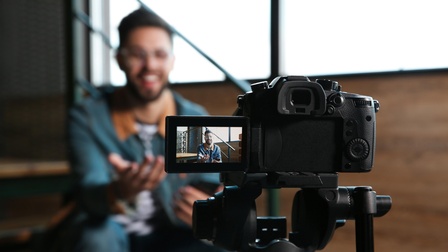 Person sitzt auf eine Stiege und nimmt ein Video von sich selbst auf, eine Kamera steht auf einem Stativ und der Fokus liegt am Livescreen-Bildschirm