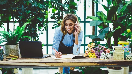 Lächelnde Person in Schürze nimmt über Tisch gebeugt Notizen vor und telefoniert mit Smartphone, ringsum Blumensträuße, Pflanzen, Papierboxen und ein Laptop