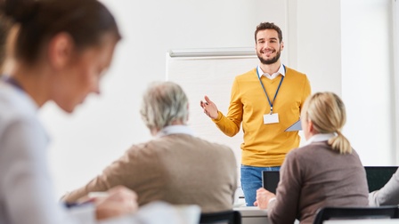 Rückenansicht von fünf Personen, die in einem Raum an Tisch sitzen. Zwei Personen haben vor sich einen aufgeklappten Laptop. Ihnen gegenüber steht eine Person, die einen Ausweis um den Hals baumeln hat und ein Klemmbrett hält.