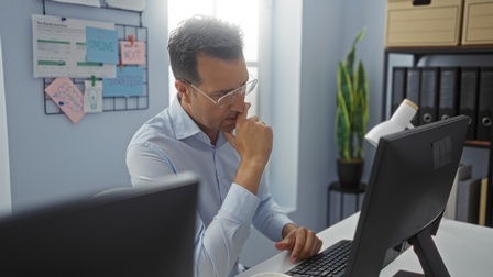 Eine Person mit Brille sitzt an einem Tisch, blickt auf einen Monitor und hält eine Hand an den Mund. Hinter der Person hängen an einer Vorrichtung verschiedene Zettel mit Stichwörtern wie Strategie.
