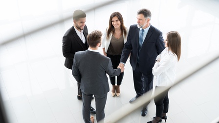Fünf Personen in Businesskleidung stehen in einem Kreis, zwei der Personen geben sich die Hand