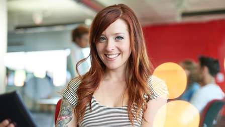 Person mit rötlichen langen Haaren trägt mit gestreiftem Shirt, Tattoo am rechten Oberarm blickt freudig in die Kamera und hält ein Tablet in der Hand, im Hintergrund  sitzen und stehen weitere Personen in einem roten Raum