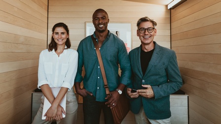 Drei lächelnde Personen stehen nebeneinander, im Hintergrund Holzwand