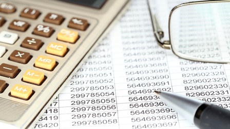 Eine Liste mit Zahlenreihen, daneben ein Taschenrechner, eine Brille und ein Kugelschreiber