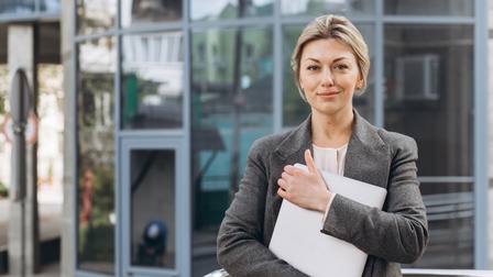 Porträt einer sanft lächelnden Person die zugeklapptes Notebook umfasst hält, im Hintergrund verschwommen Glasfassade eines Gebäudes
