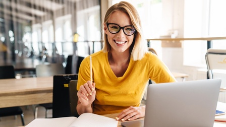 Lächelnde Person mit Brille und erhobenem Stift in der Hand sitzt vor aufgeklapptem Laptop und Büchern 
