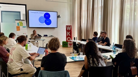 Gruppenbild des Workshops, vorne im Raum steht die Referentin, im U um sie herum sitzen die Teilnehmer:innen