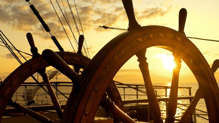 braunes Steuerrad eines Schiffes, im Hintergrund untergehende Sonne am Himmel, die durchs Steuerrad scheint