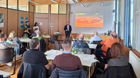 Raum mit mehreren Personen, die an Tischen sitzen, vorne steht ein Referent und hält eine Präsentation