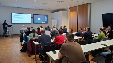 In einem Raum sitzen mehrere Menschen an Tischen und hören dem Referenten zu, der vorne steht