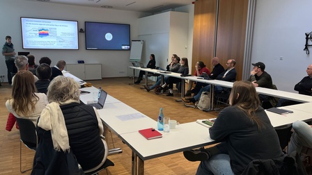 Personen sitzen in einem Seminarraum und hören dem Vortrag des Referenten zu, der vorne steht und seine Folien präsentiert