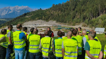 Eine Gruppe von Menschen sieht man mit dem Rücken zur Kamera, sie tragen alle eine gelbe Warnjacke mit der Aufschrift Besucher. Sie stehen im Freien und ganz hinten im Bild sieht man einen Mann von vorne, der zur Gruppe der Besucher spricht.
