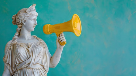 A statue of a woman holding a megaphone. The statue is yellow and white. The woman is making a loud noise, which could be interpreted as a call to action or a call for attention