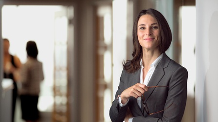 Person mit sanften Lächeln hält Brille in der Hand, im Hintergrund weitere Personen