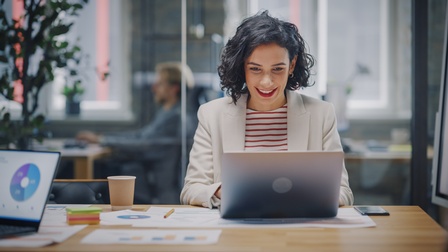 Person sitzt freudig bei einem Besprechungstisch und blickt auf einen aufgeklappten Laptop, hinter der Glaswand sitzt eine weitere Person bei einem Schreibtisch