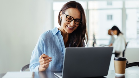 Nahaufnahme einer Person mit Brille, die vor einem aufgeklappten Laptop sitzt. Der Laptop steht auf einem dunkelbraunen Holztisch, daneben steht ein Coffee-to-go-Becher, ebenso liegen ein Notizblock und ein Smartphone auf dem Tisch