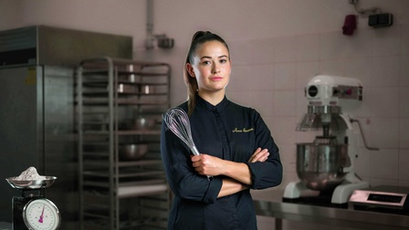 Eine Konditoren steht mit verschränkten Armen und einem Schneebesen in der Hand in der Backstube