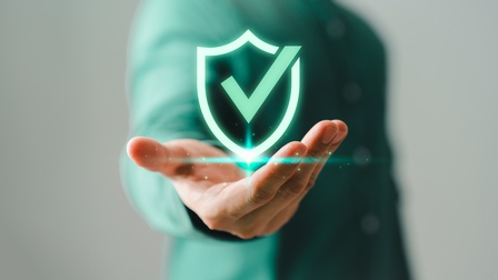 Im Vordergrund ist die Grafik eines grünen Hakens. Darunter ist die Hand einer Person