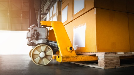 Nahaufnahme des unteren Teils eines Gabelstaplers, der eine Palette hochhebt, auf der gestapelte Kartons stehen