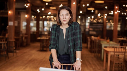 Portrait von einer Person mit schulterlangen braunen Haaren, grüner Bluse und grünem kariertem Hemd vor einem beleuchteten Restaurantambiente.