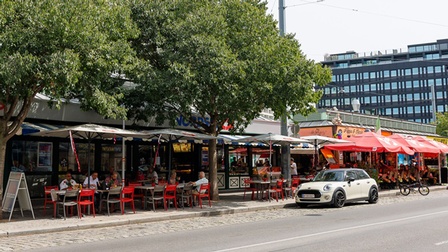 Ansicht eines Marktes von der Straßenseite aus: Verschiedene Markstände mit roten und weißen Sonnenschirmen unter denen Menschen an Tischen sitzen, davor parkt ein weißes Auto