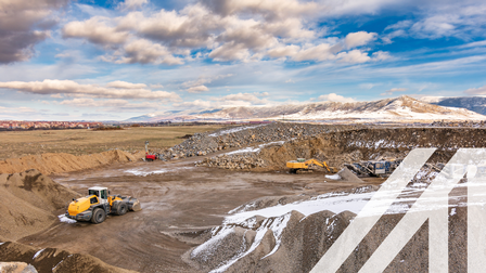Gelber Bagger, der Gestein in einem teilweise verschneiten Tagebau bewegt, Hügel aus aufgeschüttetem Bergreich, im Hintergrund verschneite Berge und bewölkter Himmel