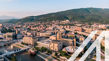 Stadtansicht Skopje: Blick von oben auf die Stadt mit historischen Gebäuden und Monumenten, Fluss Vardar mit Brücken dre Hauptstadt von Mazedonien unter blauem Himmel. Im Hintergrund sieht man eine begrünte hügelige Landschaft, den Berg Vodno.
