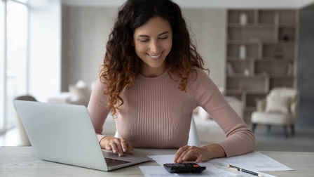 Frontalaufnahme einer Person, die hinter einem Tisch sitzt. Rechts vor der Person ist ein aufgeklappter Laptop. Links vor ihr ist ein Taschenrechner, auf den sie blickt. Unter und um den Taschenrechner sind bedruckte Zettel