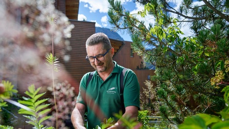 Lächelnde Person mit Brillen steht in Garten und schneidet Pflanzen, im Hintergrund verschwommen Holzfassade, ringsum grüne Pflanzen