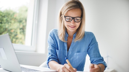 Sitzende Person mit langen blonden Haaren und Brille in blauem Hemdmacht Notizen, daneben Laptop aufgeklappt und Smartphone liegend