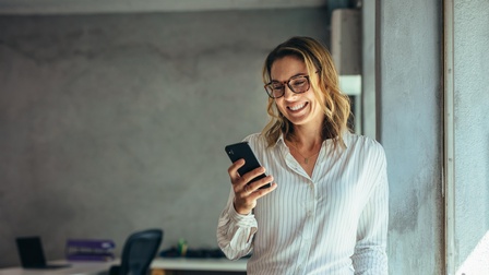Person mit Brillen blickt freudestrahlend auf Smartphone, im Hintergrund verschwommen Schreibtisch mit Sessel