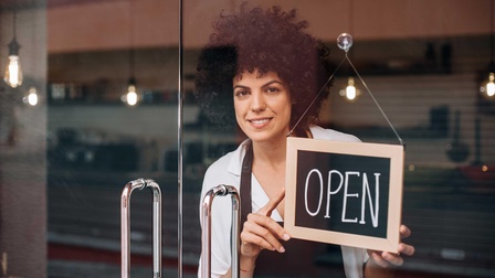 Person aus Geschäftslokal blickend, eine Tafel mit der Aufschrift 