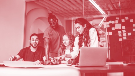 Junges fröhliches Team bestehend aus zwei Männern und zwei Frauen arbeitet gemeinsam an einem großen Glastisch mit Laptop