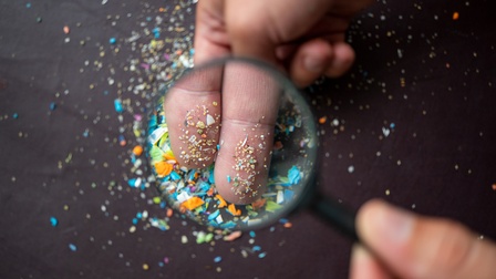 Detailansicht von zwei Fingern auf denen kleine bunte Mikroplastikpartikel liegen, ringsum weitere Partikel verstreut, darüber Vergrößerungslupe