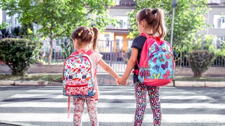 Zwei Kinder halten Hände und stehen vor einem Zebrastreifen. Dabei schauen sie nach links. Beide tragen einen Rucksack und bunte Bekleidung mit verschiedenen Muster. Im Hintergrund ist ein Haus mit Zaun, eine Wiese mit Sträuchern und Bäume.