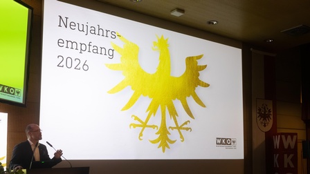Eine Person steht am Rednerpult und spricht in ein Mikrofon. Hinter der Person ist eine große Leinwand mit der Aufschrift „Neujahrsempfang 2026“ und einem großen goldenen Adler zu sehen. Im Raum sitzen und stehen mehrere Personen an Stehtischen und hören zu. Der Raum ist hell beleuchtet und als Veranstaltungssaal erkennbar.