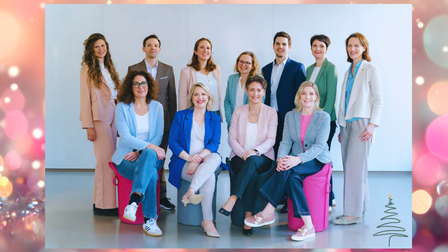 Gruppenfoto eines professionellen Teams, Frauen und Männer in Businesskleidung, stehend und sitzend in einem hellen Raum.