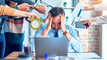 Mann hät sich die Hände an den Kopf, während Hände mit Papierkram und Uhren ihn von rechts und links unter Stress setzen