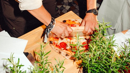 Eine Pizza wird gerade geschnitten, davor italienische Kräuter
