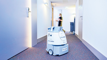 Ein Reinigungsroboter mit Dr. Schilhan-Logo, etwa halb so hoch wie ein Mensch, fährt auf einem Hotelgang.