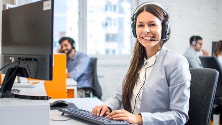 Person mit langen dunklen Haaren und Businesskleidung sitzt an einem Schreibtisch mit Headset und tippt freudig in eine Tastatur in einem Callcenter